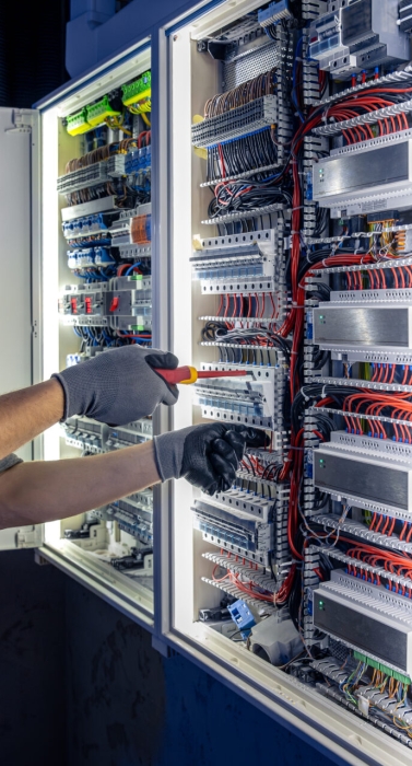 Male electrician working in switchboard with electrical connecting cable. Young adult electrical engineer in special clothes with flashlight on helmet in dark room with emergency lights in background.